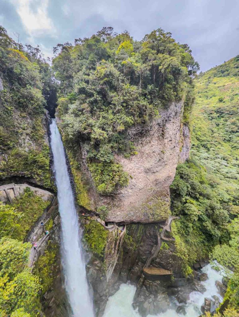 The towering Pailón del Diablo waterfall plunges down a narrow gorge, framed by lush cliffs and a winding trail with visitors exploring the site.