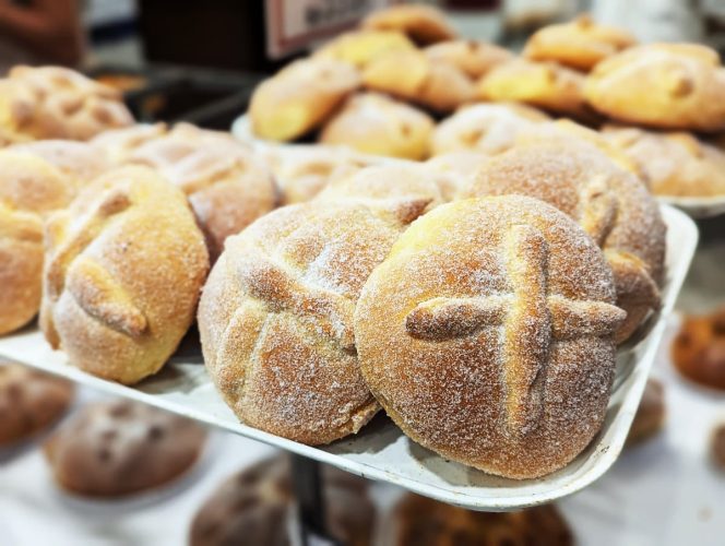 Sugar dusted pan de muerto arranged on a bakery display tray with the signature bone shaped dough decoration on top. This traditional Day of the Dead bread is commonly placed on altars and enjoyed with hot drinks.