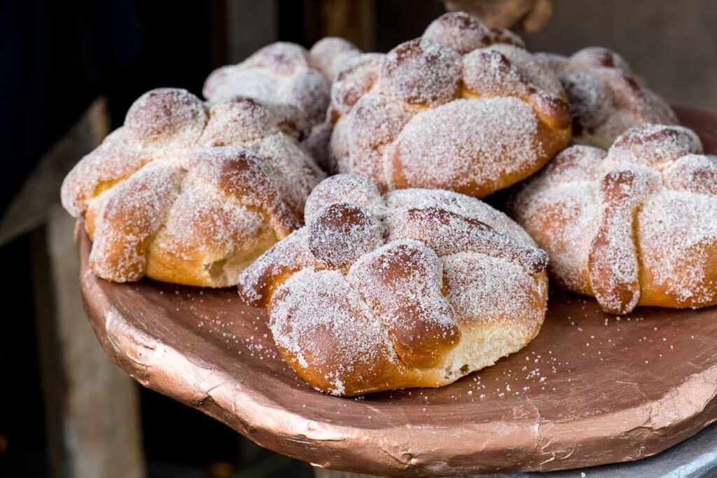 A close-up of a clay plate with six pieces of pan de muertos, a tradition for Day of the Dead in Mexico. They are decorate in the shape of bones and dusted with sugar.