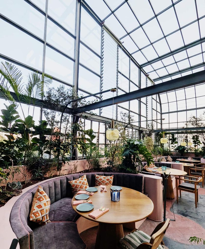 Curved velvet banquette seating and round wooden tables surrounded by greenery inside a greenhouse Mexico City rooftop.