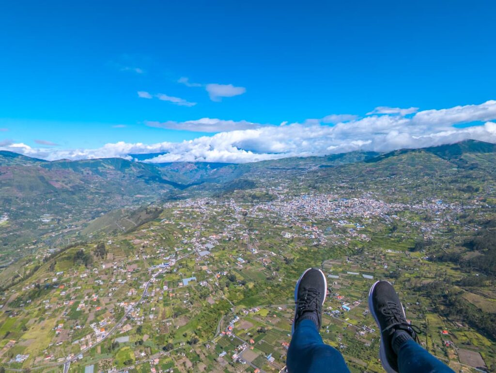 A paragliding perspective shows a person’s legs suspended high the valley near Baños Ecuador, with farmland, roads, and clusters of buildings spread out across the green valley below.