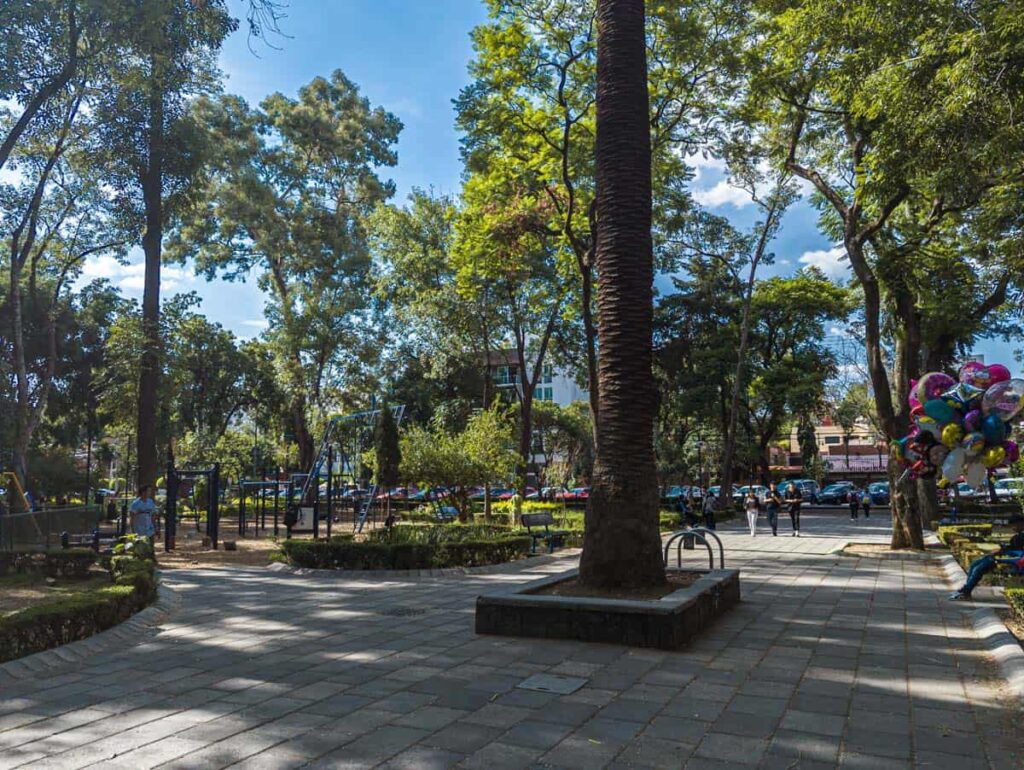 A group of three people walk along a paved pathway at Parque Arboledas in Col del Valle, CDMX. On the left is a children's play area. On the right is a man sitting on a bench holding a large bouquet of balloons for sale.