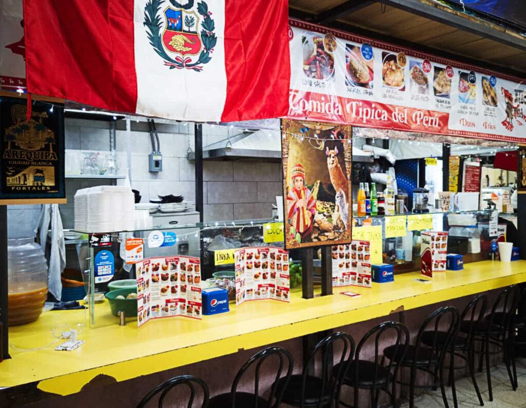 Black chairs in front of a worn yellow bar with a Peruvian flag and a banner with pictures of the Peruvian seafood available to order. Several menus and other decorations line the bar.