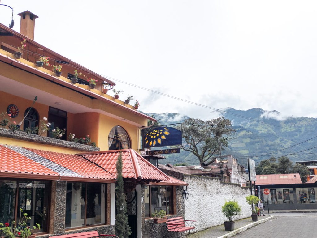 Charming exterior of La Posada del Arte, one of the best places to stay in Baños, Ecuador, with vibrant yellow walls, red-tiled roof, and flower-filled balconies, set against the misty mountains.