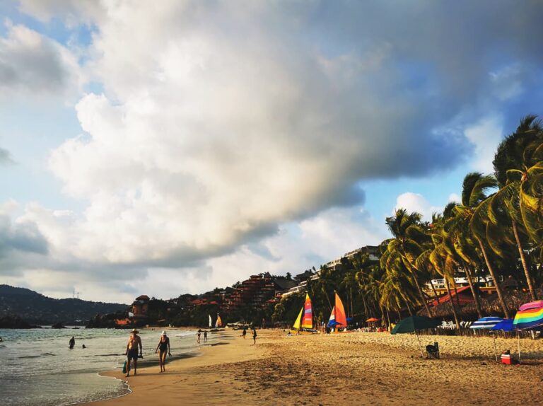 A couple wearing bathing suits walk along the waterline at Playa La Ropa in Zihuatanejo Mexico. The beach has a line of palm trees, colorful umbrellas, and several sailboats. In the background are several hotels and resorts.