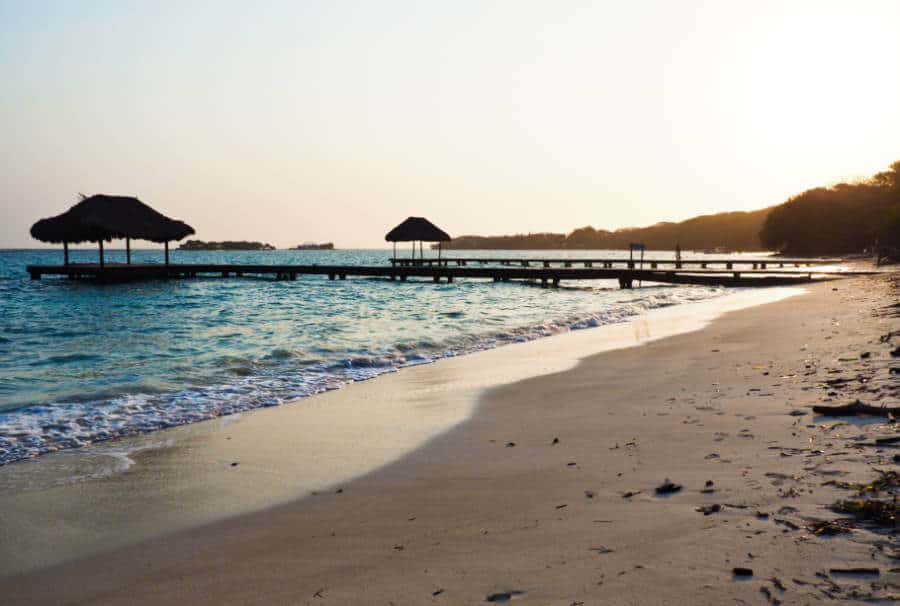Small waves of blue ocean come on to the sand at Playa Libre with long docks and palapas in the background.