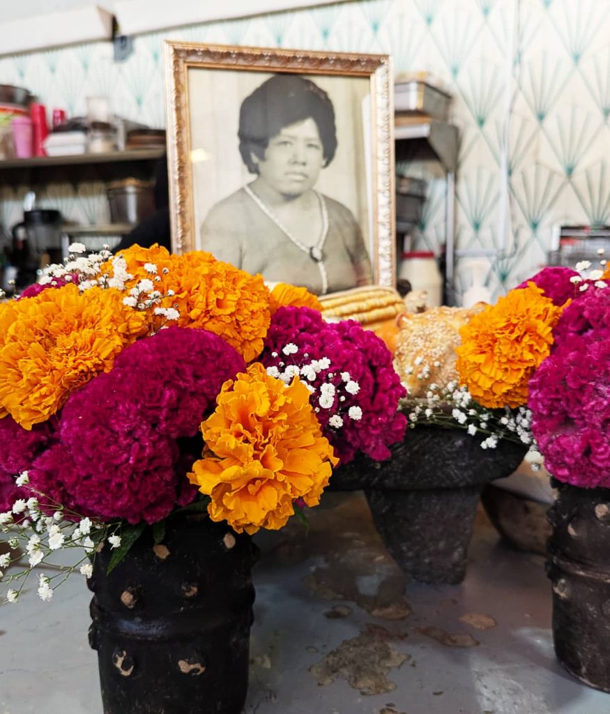 A small altar featuring a framed black and white portrait of a woman surrounded by small black clay vases filled with marigolds, cockscomb, and baby's breath flowers. As a modern addition, the portrait has become one of the most important parts of a Day of the Dead altar in Mexico.