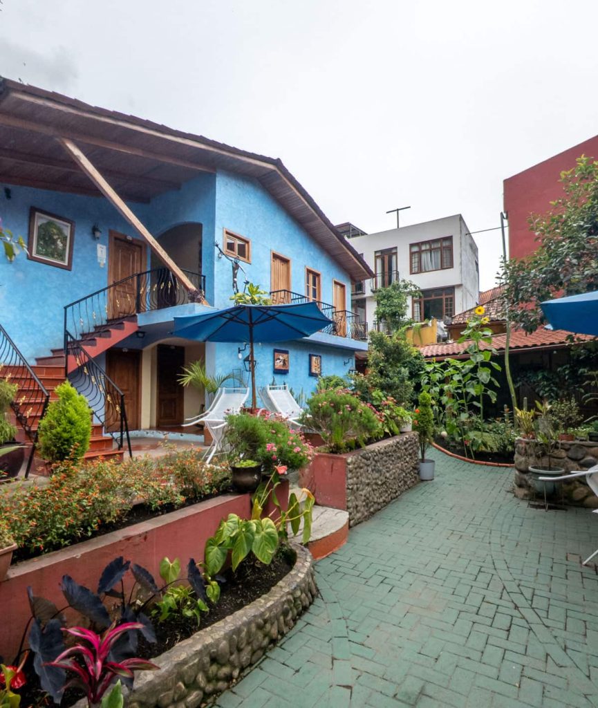 Bright courtyard garden with blooming plants, blue-painted guesthouse, and relaxing lounge chairs under umbrellas at Posada del Arte, a great option for travelers wondering where to stay in Baños, Ecuador.