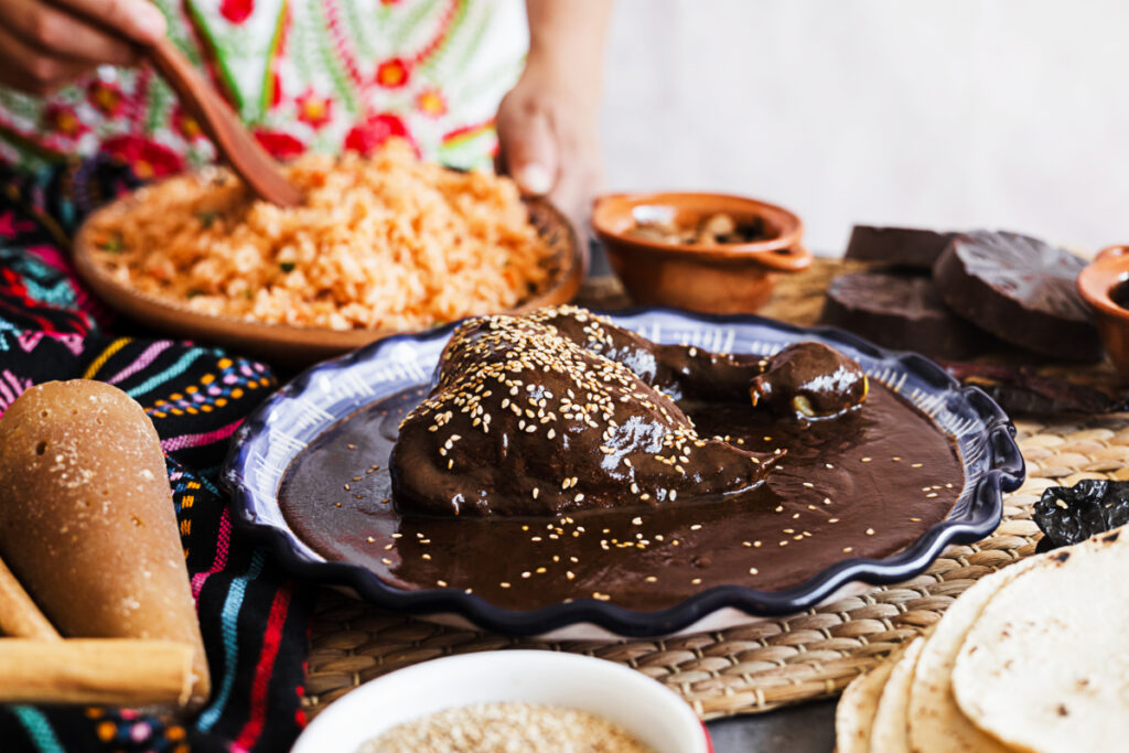 One of the most traditional Day of the Dead foods, mole negro is topped with sesame seeds on a plate. Surrounding the plate are ingredients to make mole and in the background, hands use a wooden utensil to stir a plate of rice.