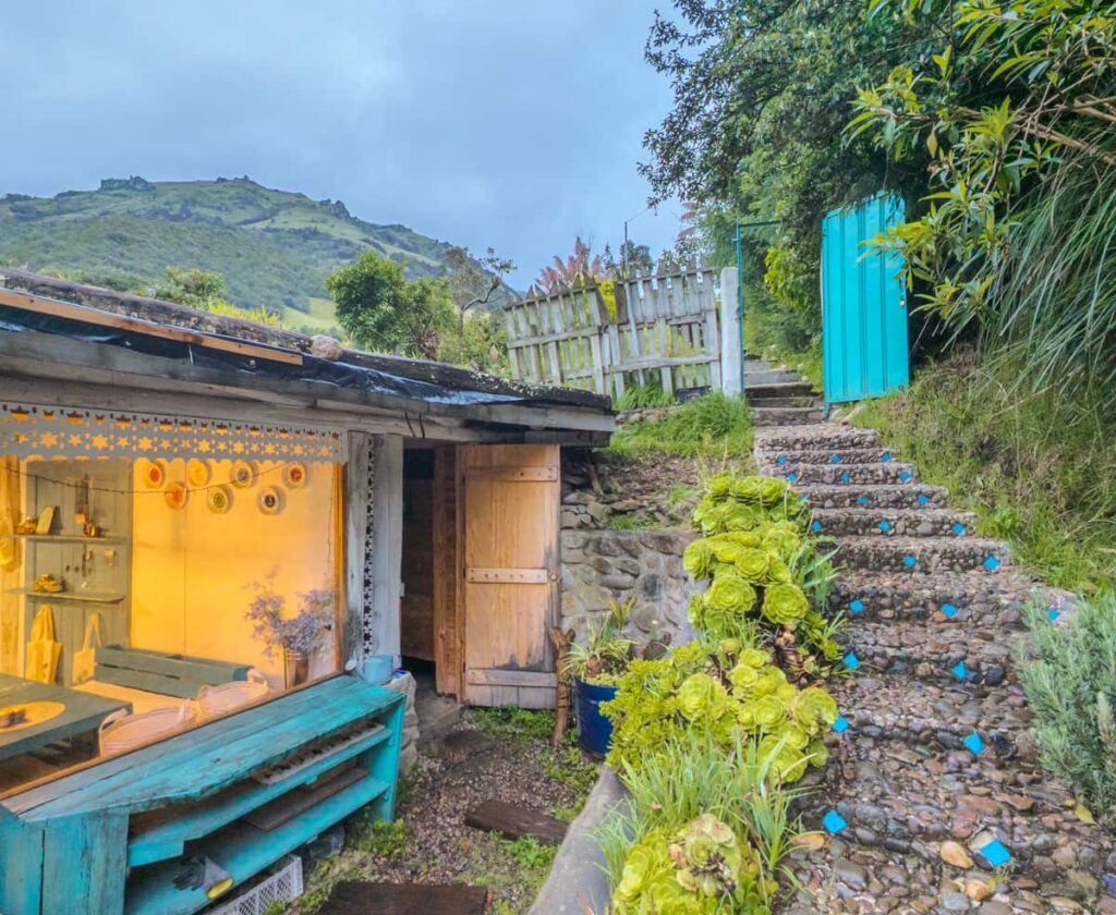 Stone steps path leading to the Pumamaqui hot springs in Cuenca. Bright green succulents line the path with a vibrant turquoise gate at the top. On the left is a small restaurant with a large glass window.