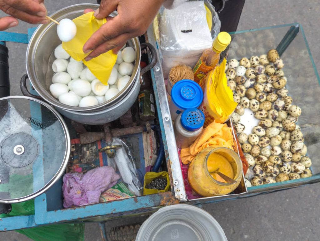 An overhead view of a street cart in Ecuador displaying a pot of peeled hard-boiled quail eggs, sauces, and seasonings, with a hand placing a skewered egg into a yellow bag. On the other side of the cart are eggs still in their shell.