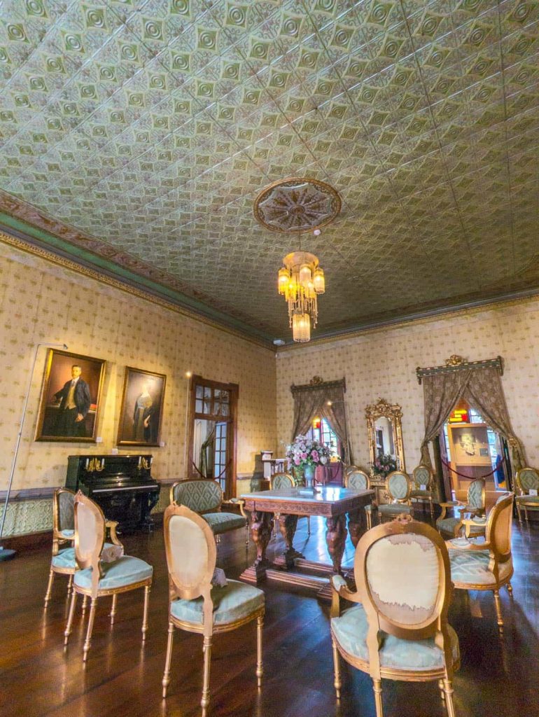 Inside the Remigio Crespo home, a historic Cuenca museum, a lavish salon features antique furniture, portraits, a piano, and a detailed green patterned ceiling.