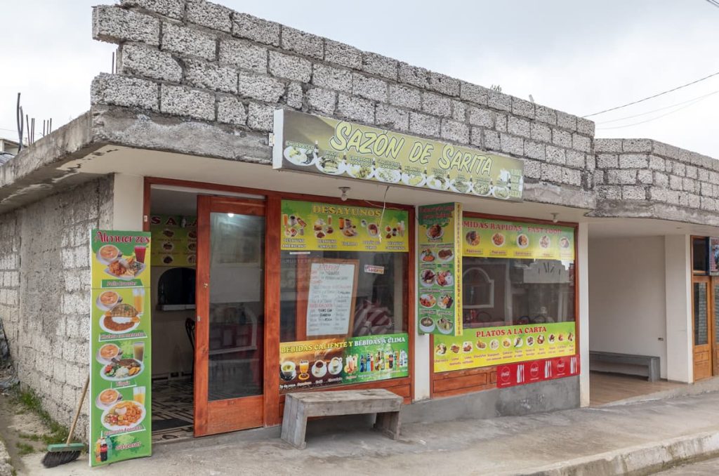 The exterior of a simple local restaurant called Sazón de Sarita in Quilotoa Ecuador, with colorful signs advertising typical Ecuadorian food.