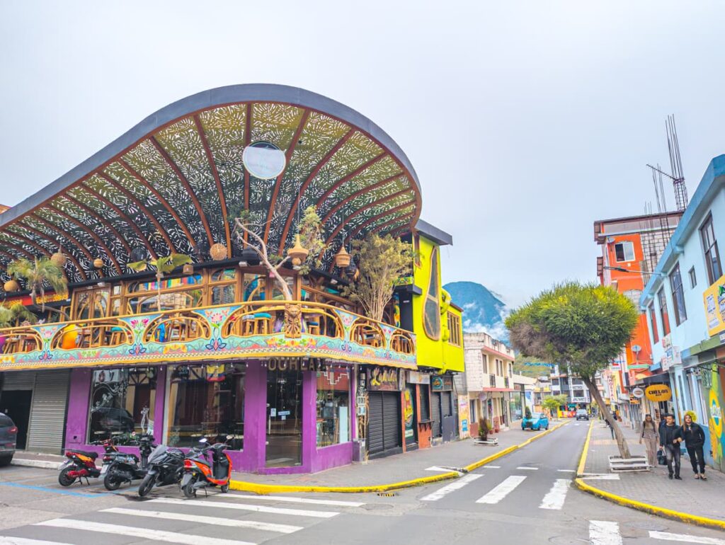 A vibrant street scene in Baños Ecuador features a uniquely designed building with a curved rooftop decorated with hanging plants and intricate patterns, surrounded by shops and scooters.