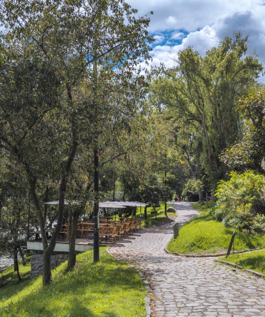 A cobblestone path winds through shaded greenery along the Río Tomebamba in Cuenca Ecuador, with outdoor seating under umbrellas by the water. Strolling along this peaceful riverside is one of the most relaxing things to do in Cuenca.