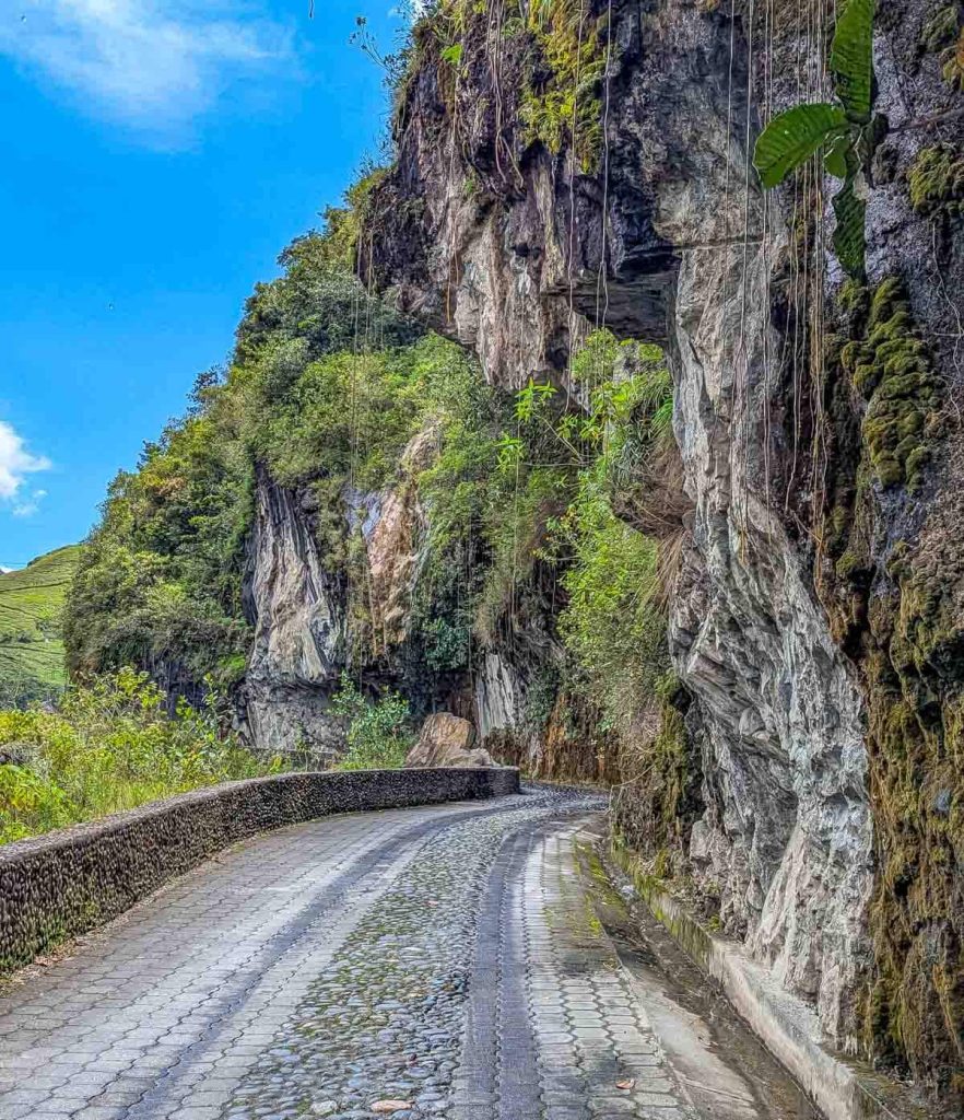 A narrow cobblestone road curves alongside a towering cliff face covered in vines and moss, one of the scenic routes on the Baños waterfall tour.