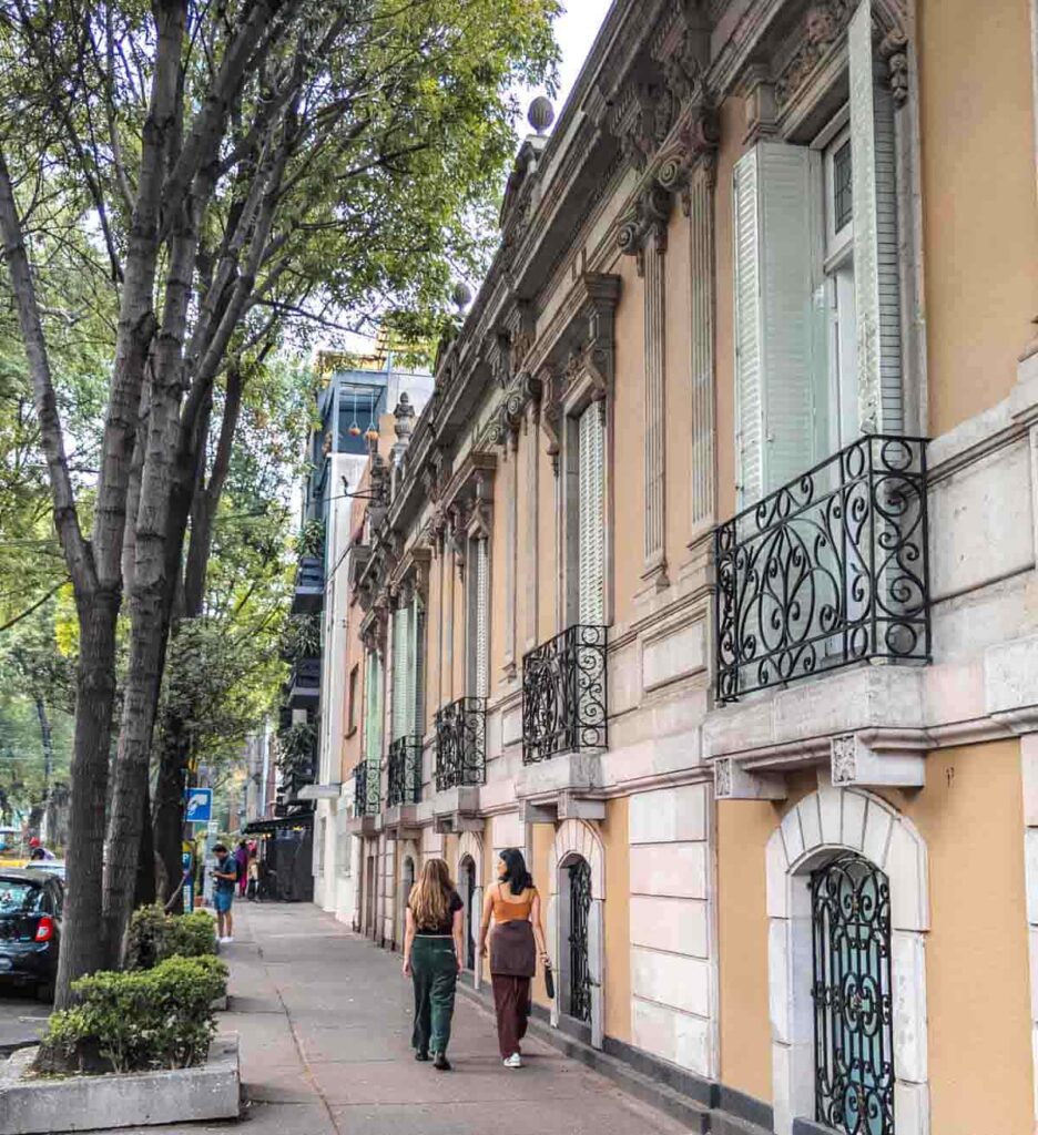Two tourists walk away from the camera along a sidewalk lined with beautiful buildings in Roma Norte, one of the best areas to stay in Mexico City. The front of the building features crown molding and iron work.