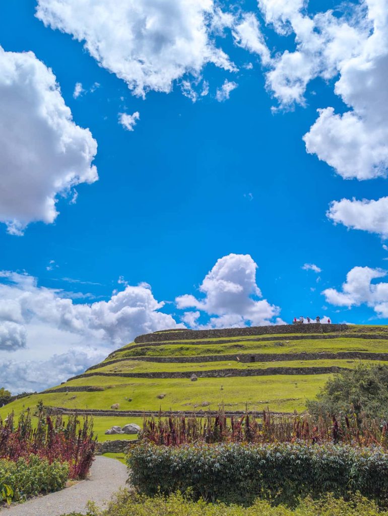Wide view of the tiered grassy terraces of the Pumapungo ruins in Cuenca with groups of visitors walking along the top.