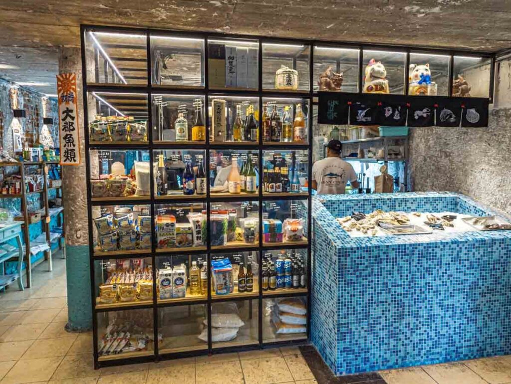 Interior of Sakanaya Japanese Restaurant in Mexico City features glass displays with Japanes food items for sale items. On the right is a blue tiled ice box with fresh fish.