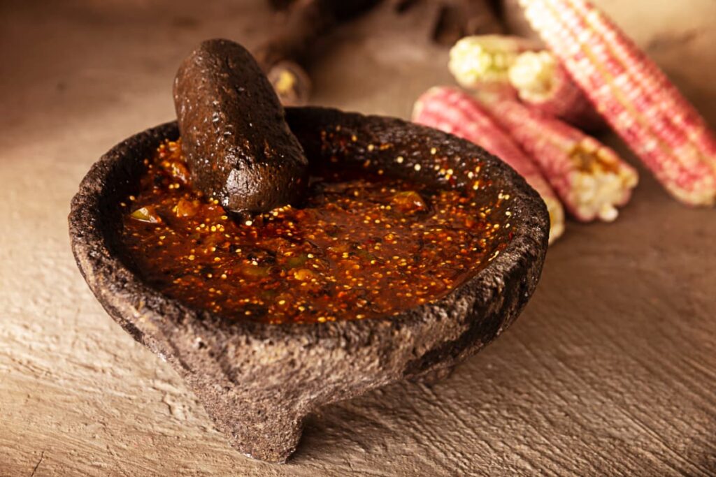 At a salsa making class in Mexico City, a stone molcajete filled with red salsa sits on a concrete surface. Behind the mortar and pestle are several ears of red corn.