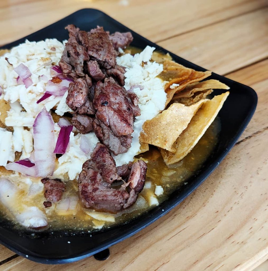 A square black plate of chilaquiles topped with salsa morita, arrachera, queso, and red onion sitting on a wooden table at Divino Chilaquiles in Mexico City.