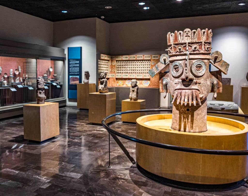 A large clay head sculpture on display in the middle of a room at the Anthropology Museum in Mexico City surrounded by smaller sculptures.