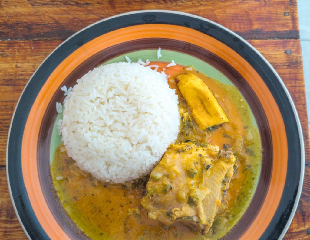 A plate of seco de pollo features a stewed chicken thigh smothered in a light brown sauce, served with a mound of white rice and a slice of ripe plantain. This classic seco is one of the most popular dishes in Ecuador.