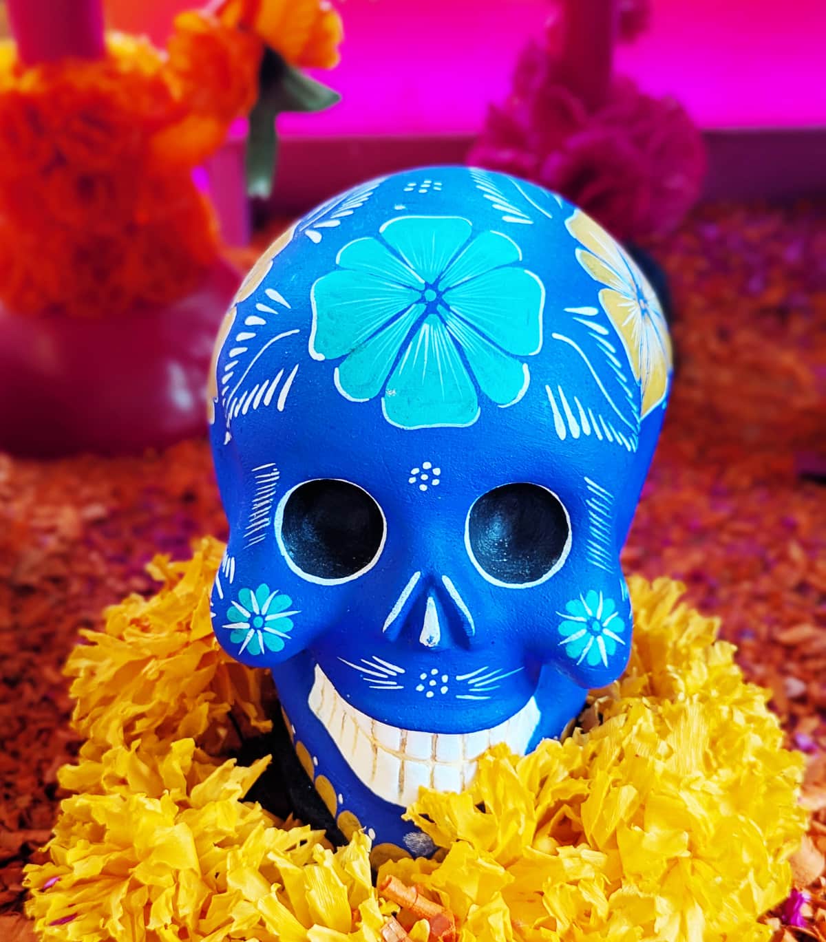 Bright blue decorative skull resting on a bed of orange marigold petals with floral patterns painted across its surface. The skull is one of the most common Day of the Dead symbols.