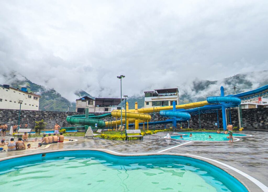 One of the hot springs in Baños Ecuador features colorful water slides and multiple pools, set against a cloudy mountain backdrop. Visitors relax in the warm mineral water.