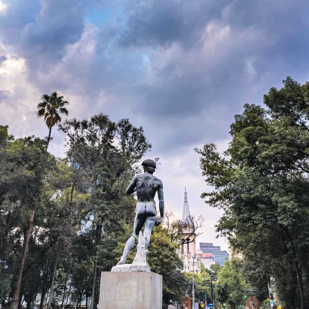 A statue of the backside of a naked man in Plaza Rio de Janeiro, a small park in the Roma Norte neighborhood of CDMX. In the background are some trees and buildings.