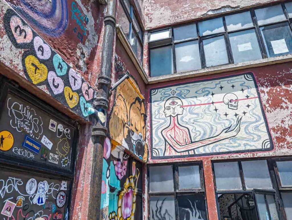 Multiple street art murals at the Toy Museum in Mexico City. The main on features a double head with third eye and a body with red veins running through. A red beam shoots out fom one of the eyes towards a skull on the right side.