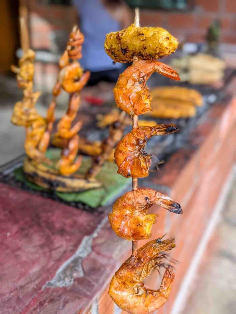 Grilled shrimp skewer with a piece of charred plantain on top, displayed in front of a barbecue setup at an Ecuadorian street food stall.