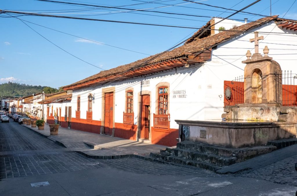 Sunlight casts warm shadows on the cobblestone streets of Pátzcuaro, Michoacán, highlighting colonial architecture and a rustic stone fountain.