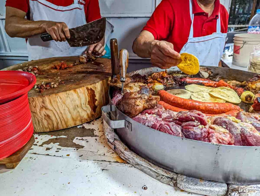 On the left a man chops and prepares suadero tacos. On the right a man heats small corn tortillas in the center of a round metal container. Surrounding the tortillas longaniza sausage, beef, and onions are cooking.