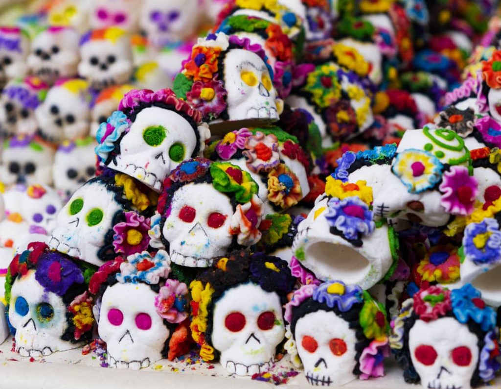 Dozens of small white sugar skulls, a Day of the Dead tradition, piled high. Each skull is decorated with bright icing flowers and colorful eye sockets in pink, green, red, and blue — ready for a Día de Muertos altar.