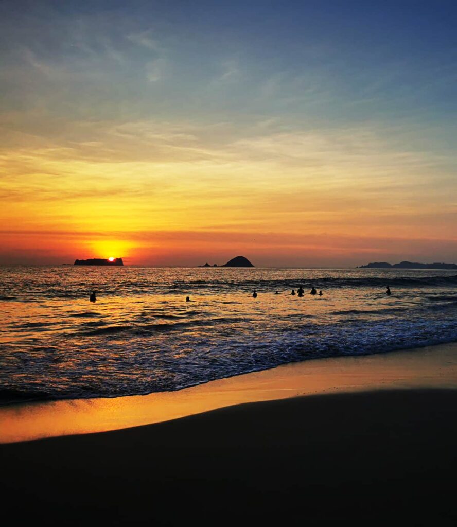 Several people play in the waves during sunset on Playa el Palmar in Ixtapa Zihuatanejo. The golden orange hues reflect into the sky and the ocean below.