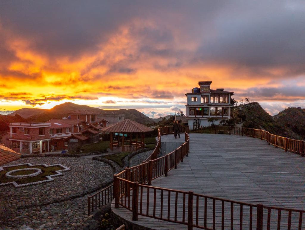 Sunset over the village near Quilotoa Lagoon, with orange and pink clouds lighting up the sky behind mountain silhouettes. A wooden boardwalk and Quilotoa Lake lookout leads to a multi-story building with large windows.