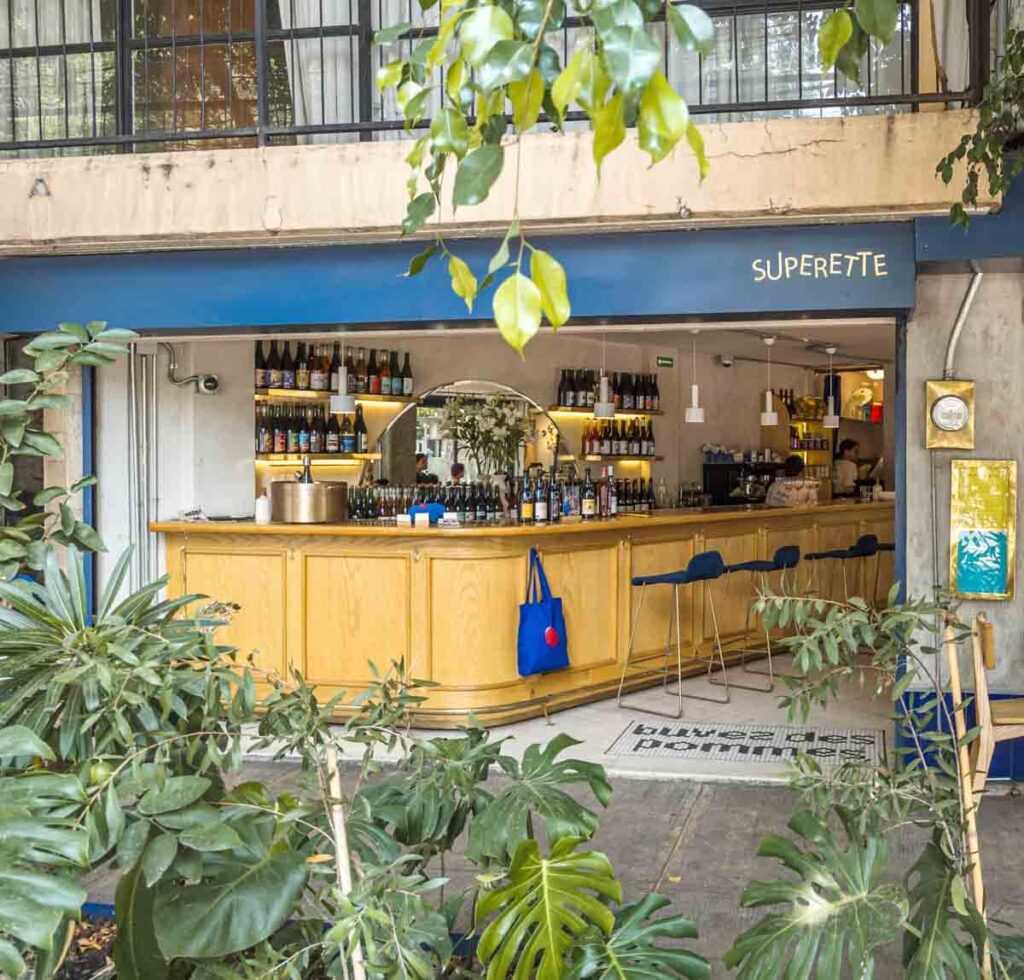 An exterior shot of Superette Restaurant bar area filled with wine bottles and liquors. In the foreground are several leafy green plants