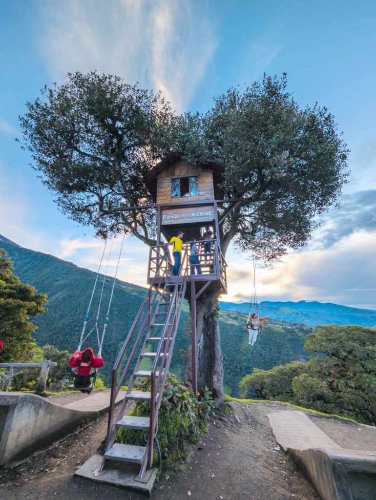A rustic treehouse sits perched in a large tree at Casa del Árbol in Baños Ecuador, with visitors swinging out over the edge overlooking a dramatic green valley below.