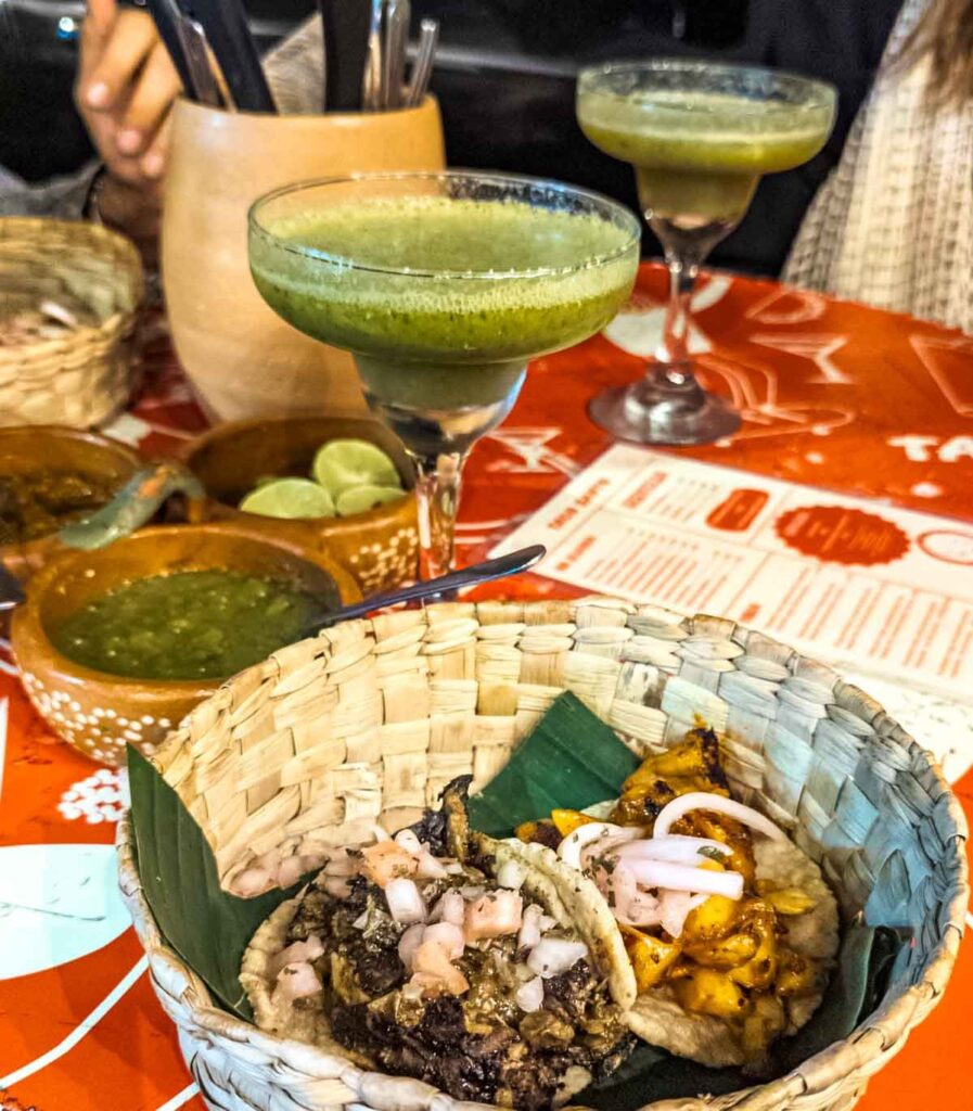 Two vegan tacos served in a woven basket lined with a banana leaf at Taco Santo Vegano in Mexico City. In the background are green jalapeno cocktails and bowls of salsa and limes.