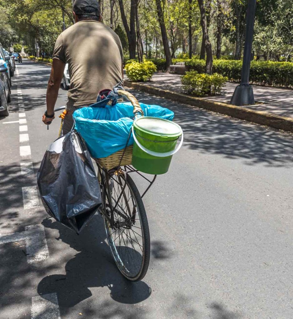 The backview of a man on his bicycle selling tacos de canasta in Mexico City. The large basket on the back of his bike is lined with blue plastic. A green tub of salsa hangs from the basket and a trash bag hangs from the left side of the bike.