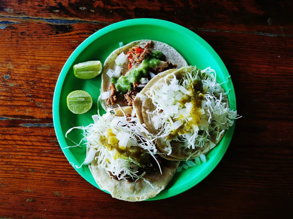 A vibrant green plate holds three traditional Mexican tacos in Ixtapa. They are generously topped with shredded cabbage and salsa. Accompanied by fresh lime wedges, they sit on a rustic wooden table.