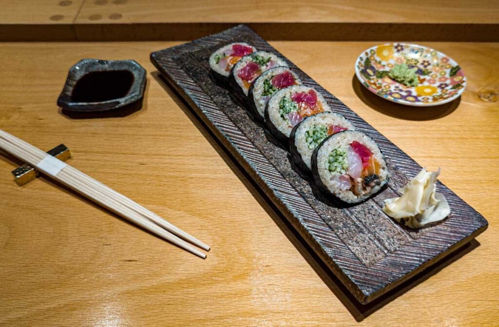 Colorful sushi roll in Mexico City displayed on a rectangle plate with ginger. Nearby are chopsticks, a small dish of wasabi, and a dish of soy sauce.
