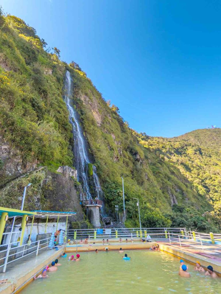 Visitors soak in warm yellowish thermal pools at Termas de la Virgen, located at the base of a tall waterfall cascading down a steep cliffside in Baños Ecuador.