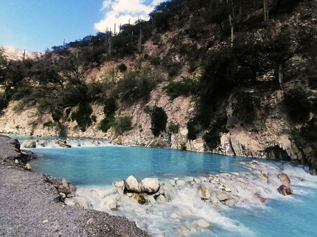 The bright blue thermal river at Grutas Tolantongo is stepped with rocks to create small pools. In the background is a rockface with cactus and other plants growing.