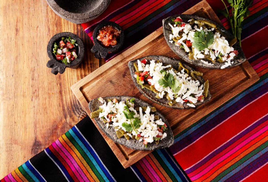 Overhead view of a table dressed with colorful Mexican textiles and a cutting board with three tlacoyos made during a cooking class in Mexico City.