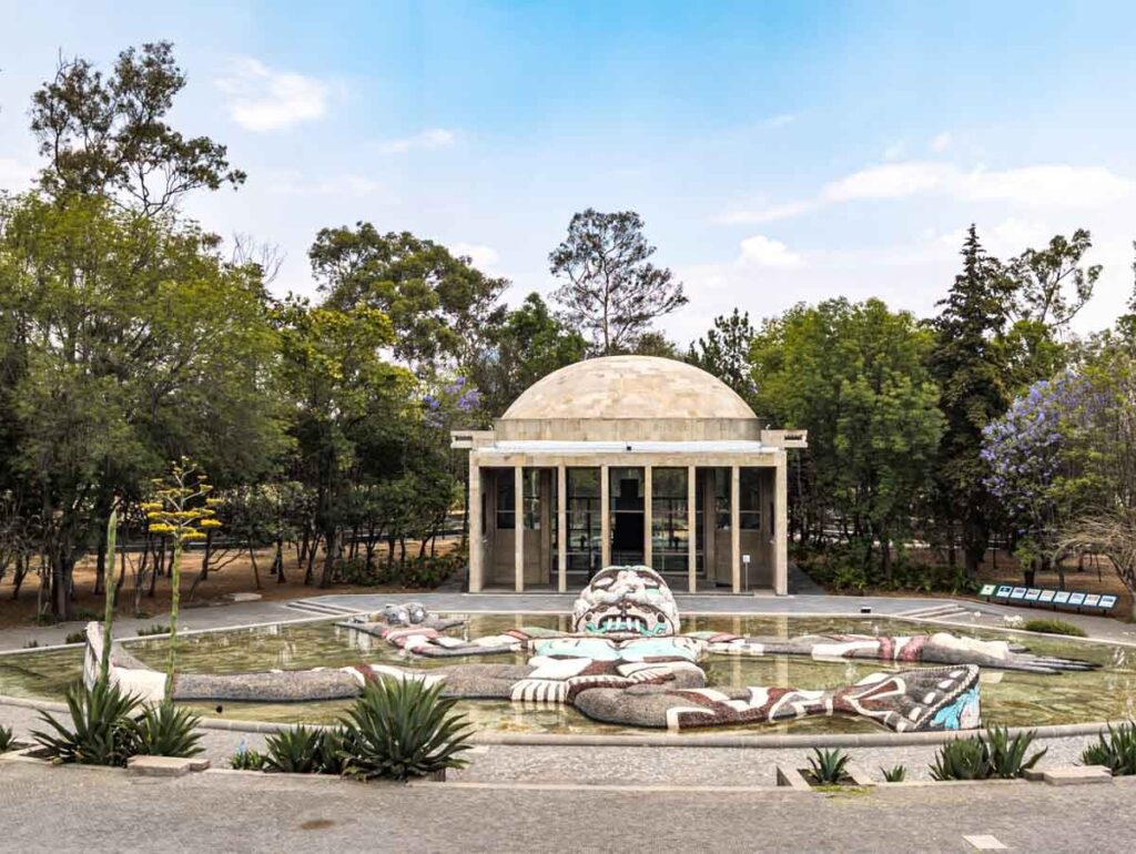 Surrounded by trees and the museum building, the Tlaloc rain god sculpture lays flat in a shallow pool of water at the Museo del Cárcamo de Dolores in Mexico City.