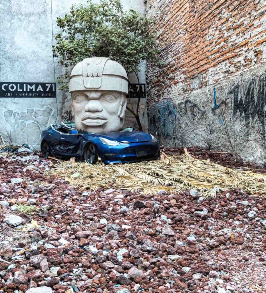 A concrete replica of a Tolmec head sits on top of a crushed blue Tesla in an empty lot in Mexico City.