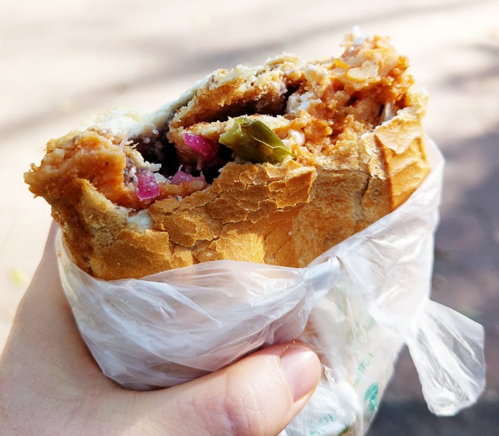 A hand holding a torta de chilaquiles in Mexico City. It's wrapped in a plastic bag, stuffed with crispy breaded chicken, salsa, crema, and onion.