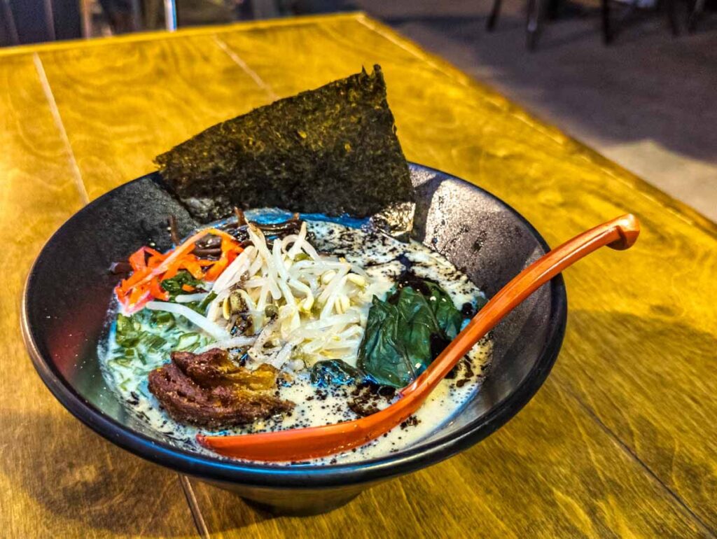 A bowl of vegan ramen in Mexico City is served in a black bowl with large orange spoon and a square piece of nori seaweed. The ramen contains a mix of noodles and veggies.
